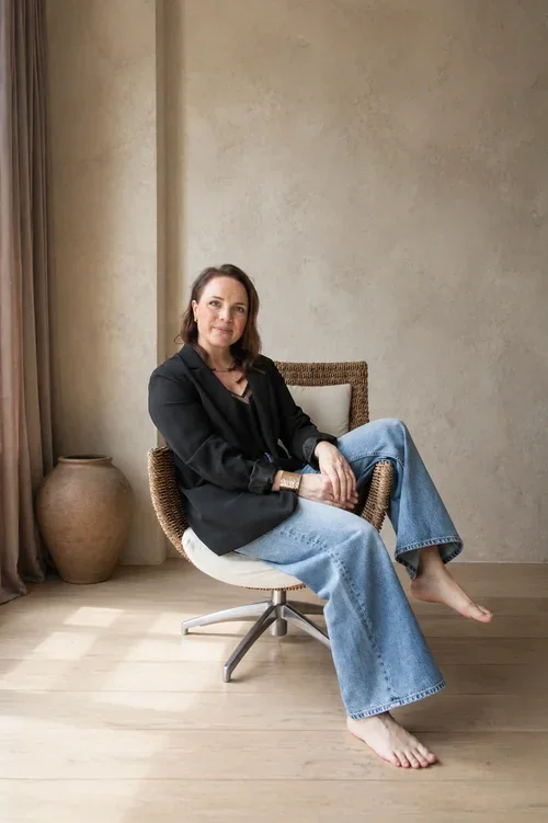 Kvinne med mørkt hår, iført svart blazer og blå jeans, som sitter avslappet i en flettet kurvstol og ser rolig mot kameraet.