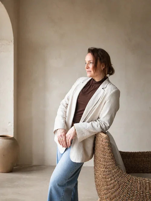 Kvinne i hvit blazer og brun topp som står i profil og lener seg mot ryggen av en flettet stol i et lyst rom med kalkede vegger.