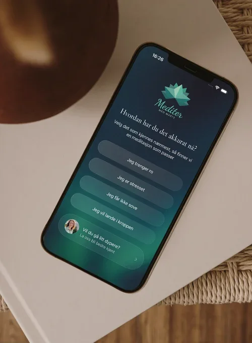 Bildet av mobil som ligger på en bok på et bord. Mobilen viser innholdet i appen Meditere med Marte.